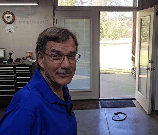 Man in a blue polo standing in a workshop with an open door, tools in the background, and a rubber snake on the floor—an effective bird deterrent trick